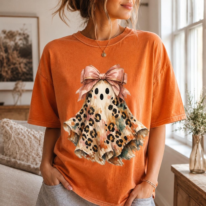 May include: An orange short-sleeve t-shirt featuring a central graphic of a ghost adorned with a large pink bow. The ghost has a beige body with a leopard print pattern and colorful brushstroke accents, including black eyes and a small mouth.