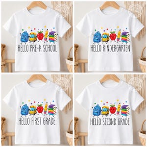 Puede incluir: Una camiseta blanca con un gráfico colorido de una mochila azul, un autobús escolar amarillo, una manzana roja, un lápiz amarillo y una pila de libros con caras sonrientes, rodeados de estrellas y el texto Hello Pre-K School, Kindergarten, First Grade o Second Grade.