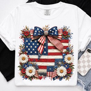 Peut inclure: T-shirt blanc avec un motif patriotique. Le motif comprend un drapeau américain en patchwork avec un nœud bleu orné d'étoiles blanches et de rayures rouges. Le drapeau est entouré de marguerites, de fleurs rouges et d'accents bleus.