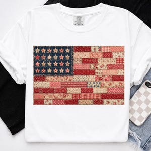 May include: White t-shirt featuring a patchwork American flag design. The flag incorporates various red, white, and blue patterned fabrics, including floral and checkered patterns. The shirt has a crew neck and short sleeves, with the brand name "Comfort Colors" visible.
