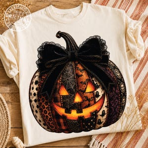 May include: A cream-colored t-shirt featuring a large graphic of a jack-o'-lantern pumpkin. The pumpkin is decorated with a black lace bow and various patterns including spiderwebs, bats, ghosts, skulls, leopard print, and plaid.