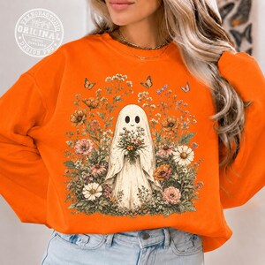Pu&ograve; includere: Una felpa arancione con una grafica centrale raffigurante un fantasma bianco che tiene un bouquet di fiori, circondato da una fitta disposizione di fiori di campo colorati e farfalle. Il design include vari fiori nelle tonalit&agrave; rosa, arancione e bianco.