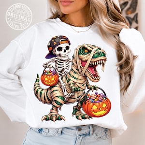 May include: A white long-sleeve sweatshirt featuring a graphic of a skeleton wearing a baseball cap and riding a mummy-wrapped Tyrannosaurus Rex. Both figures hold orange jack-o'-lantern buckets filled with colorful candy for Halloween.