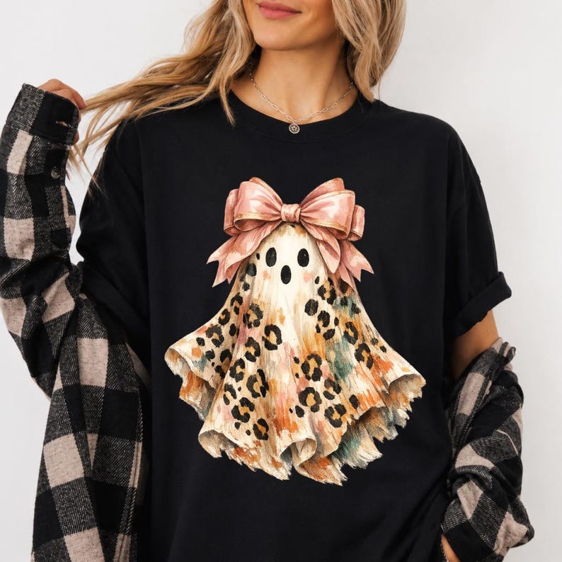 May include: A black t-shirt featuring a graphic of a ghost wearing a large pink bow. The ghost has a tan and brown leopard print pattern with watercolor accents in orange and teal. The shirt is worn with a black and tan plaid flannel shirt.