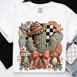 May include: A white t-shirt featuring a central graphic of a green cactus wearing a leopard print cowboy hat. The design includes a large orange floral bow, two horseshoes, stars, and a patchwork background of checkered and paisley patterns.