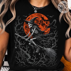 May include: A black t-shirt featuring a graphic of a skeleton wearing a witch hat and riding a broomstick against a large orange moon. The design includes silhouettes of flying bats, ravens, bare tree branches, and spider webs in the background.