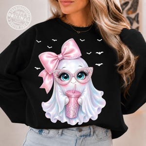 May include: A black sweatshirt featuring a graphic of a white ghost wearing pink glittery glasses and a large pink bow. The ghost holds a pink glittery tumbler cup, surrounded by small white bat silhouettes against the black fabric background.