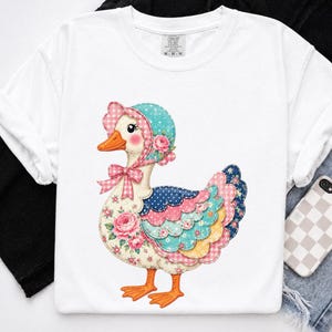 May include: A white t-shirt featuring a graphic of a duck wearing a bonnet. The duck has a floral pattern on its body, a pink gingham bow, and a bonnet with pink roses. The wings are layered with blue, pink, and yellow polka dot and floral fabrics.