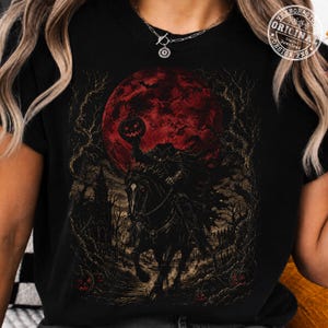 May include: A black t-shirt featuring a graphic print of a headless horseman riding a horse under a large red moon. The scene includes dark, bare trees, a silhouette of a building, and small pumpkins on the ground. The print has a dark, detailed style.
