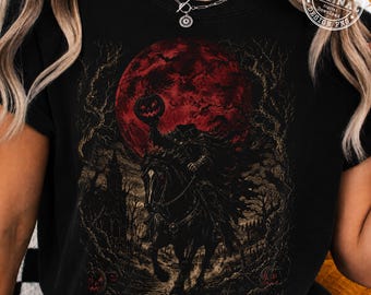 May include: A black t-shirt featuring a graphic print of a headless horseman riding a horse under a large red moon. The scene includes dark, bare trees, a silhouette of a building, and small pumpkins on the ground. The print has a dark, detailed style.