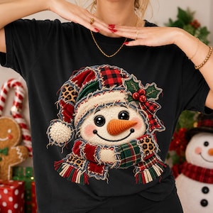 Pode incluir: Uma t-shirt preta de manga curta com um gráfico central de um boneco de neve com um gorro e cachecol de retalhos. O boneco de neve tem um rosto branco texturizado, um nariz de cenoura laranja e é decorado com bagas vermelhas e azevinho.