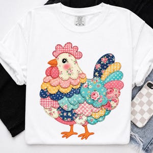 以下が含まれることがあります： 白のTシャツで、中央に様々なカラフルな柄の布地パッチで構成された鶏のグラフィックが描かれています。鶏のデザインには、ピンク、青、黄色、赤の色合いで、花柄、水玉模様、ギンガムチェックのプリントが含まれています。
