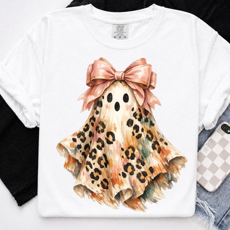 May include: A white short-sleeved t-shirt featuring a central graphic of a ghost wearing a large, light pink bow. The ghost has a tan and orange watercolor-style body covered in black leopard print spots, with two black eyes and an open mouth.
