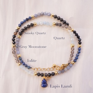 Może przedstawiać: Wielowarstwowy naszyjnik z koralikami z kwarcem dymnym, szarym kamieniem księżycowym, iolitem i lapis lazuli. Naszyjnik ma złote akcenty i niebieską zawieszkę z lapis lazuli. Tekst na zdjęciu brzmi "Smoky Quartz", "Quartz", "Gray Moonstone", "Iolite" i "Lapis Lazuli".