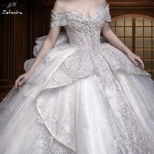 Op de afbeelding: Een witte off-the-shoulder baljurk met ingewikkelde zilveren kralen en borduursels. Het lijfje is getailleerd en de rok is volumineus met gelaagde tule. De jurk heeft een sweetheart halslijn en korte mouwen. De merknaam "Zelmiria" is zichtbaar.