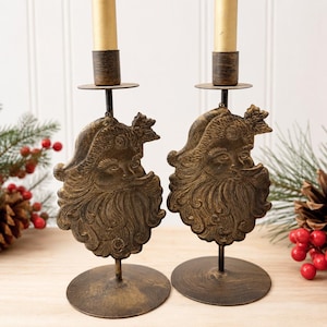 Könnte beinhalten: Zwei passende Kerzenhalter aus Metall mit einem dreidimensionalen, antikgoldfarbenen Weihnachtsmann-Gesichtsmotiv. Jeder Halter verfügt über einen runden Fuß, einen dünnen vertikalen Stab und eine obere Tülle für eine goldene Stabkerze.