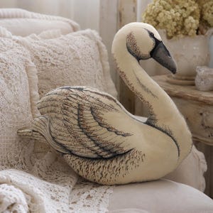 May include: A plush swan-shaped cushion featuring a cream-coloured fabric base with detailed black and brown ink-style illustrations of feathers and wings. The swan has a curved neck, a dark beak, and a soft, stuffed body designed in a sitting pose.