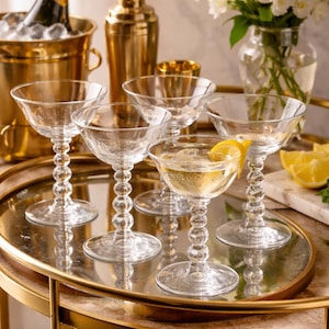 May include: A bar cart with champagne glasses, a bottle of champagne in an ice bucket, and a cocktail shaker. One glass has a lemon slice. The glasses have a beaded stem design.
