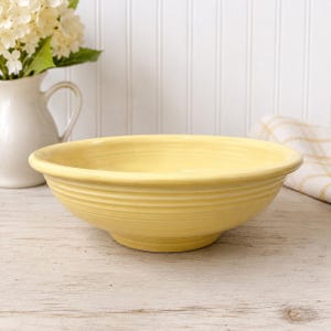 Puede incluir: Un cuenco de cerámica amarillo pálido con un borde ancho y un exterior acanalado. El cuenco está sobre una superficie de madera de color claro. Una jarra blanca con flores está en el fondo.