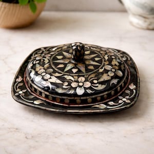 May include: A square, black decorative box with a lid, decorated with inlaid mother-of-pearl floral and geometric patterns. The lid has a small, dark knob. The box is on a marble surface, highlighting its intricate craftsmanship.