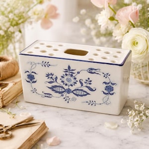 Puede incluir: Jarrón de flores rectangular de cerámica blanca con diseño floral azul. El jarrón tiene múltiples agujeros en la parte superior para colocar flores y una abertura rectangular. El jarrón está decorado con patrones florales azules.