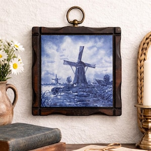 Può includere: Piastrella in ceramica incorniciata con un paesaggio di mulini a vento blu e bianco. La cornice in legno marrone scuro ha un design intagliato e un anello in ottone per appenderla. L'opera raffigura mulini, acqua e cielo nuvoloso.