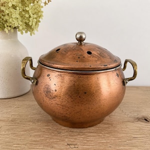 Puede incluir: Una olla redonda de cobre con una tapa abovedada a juego que presenta pequeños orificios de ventilación y un pomo central de metal. La olla tiene dos asas curvas de latón en los lados, mostrando una pátina desgastada y manchas oscuras.