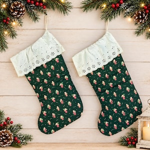 Puede incluir: Dos calcetines navideños de tela verde oscuro con un patrón repetitivo de figuras de Papá Noel. Cada calcetín tiene un puño de encaje blanco con ojales en la parte superior y un lazo para colgar. Se muestran sobre un fondo de madera clara.