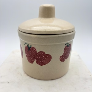 May include: A cylindrical ceramic canister with a matching lid, featuring a light beige speckled finish and a hand-painted pattern of red strawberries with green stems encircling the exterior body of the container.