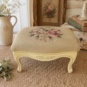 May include: A rectangular footstool with a beige needlepoint top featuring a central floral motif in shades of pink, green, and black. The stool has four curved, light cream-colored wooden legs with carved leaf details on the front apron.