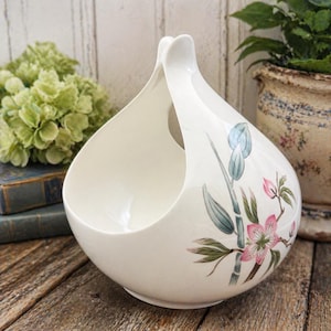 May include: A white ceramic basket with a handle, featuring a floral design with pink blossoms and green leaves. The basket has a rounded base and a wide opening, perfect for displaying items.