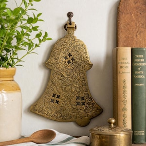 May include: A decorative brass bell, featuring detailed floral engravings and four cross-shaped apertures. It hangs from a dark metal hook. The image also shows vintage books and a wooden spoon, creating a rustic, antique aesthetic.