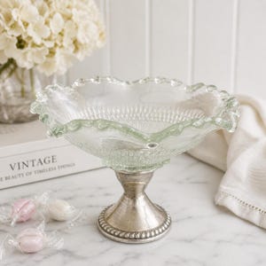 May include: A clear glass pedestal bowl with a ruffled edge and a silver-colored base. The bowl has a textured design and sits on a decorative, ornate base. The bowl is likely used for serving or display.