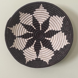 Könnte beinhalten: Ein runder, flacher geflochtener Korb mit einem kontrastierenden geometrischen Muster in Schwarz und Off-White. Das Design strahlt von der Mitte aus in einer spiralförmigen Konstruktion und erzeugt eine Reihe sich wiederholender Rautenformen.