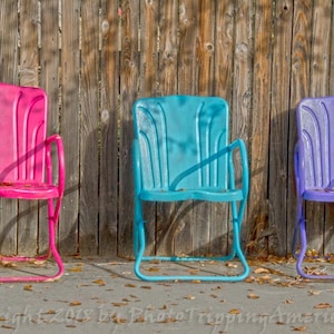 May include: Three vintage metal chairs in pink, blue, and purple, lined up in front of a wooden fence. The chairs have a simple design with a curved back and arms.