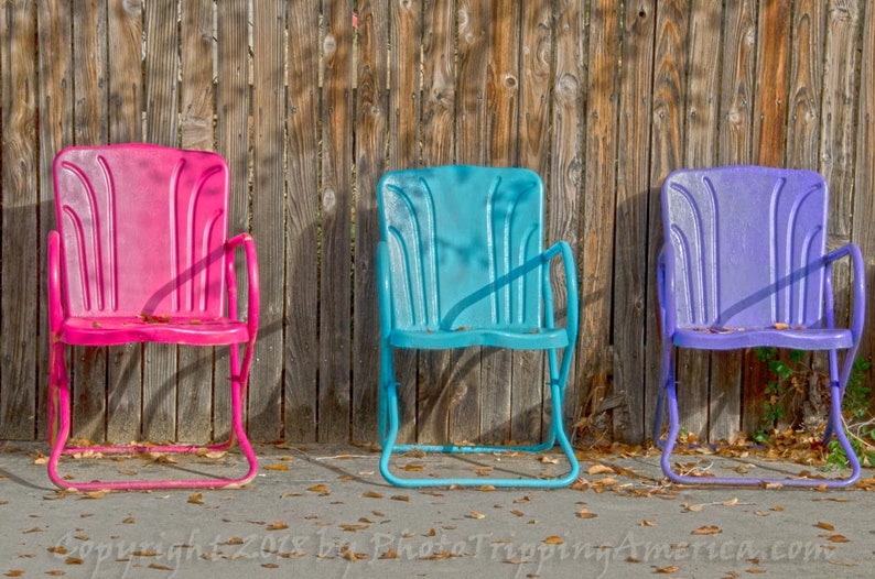 May include: Three vintage metal chairs in pink, blue, and purple, lined up in front of a wooden fence. The chairs have a simple design with a curved back and arms.