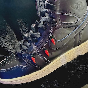 May include: Black high-top trainer with a white sole. The trainer features a silver chain with three red chilli pepper charms. The shoe has black laces and a perforated toe box.