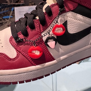 May include: A red, white, and black high-top trainer with black laces. The trainer features a black Nike swoosh and is decorated with a chain of three lip charms in red, black, and pink.