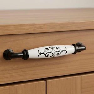Könnte beinhalten: Ein Schrankgriff mit weißer Keramikmitte und schwarzen Metallbeschlägen. Die weiße Mitte hat ein schwarzes, wirbelndes Design. Montiert an einem hellbraunen Holzschrank.