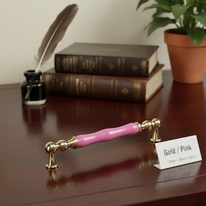 Op de afbeelding: Een gouden en roze kastgreep. De greep heeft een roze midden en gouden accenten. Het label luidt "Gold / Pink" en vermeldt afmetingen van 192 mm - 360 mm / 128 mm.