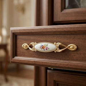 Può includere: Primo piano di un cassetto in legno con una maniglia decorativa. La maniglia è dorata con un centro ovale in ceramica bianca con un motivo floreale nei toni del rosa, dell'arancione e del viola. Il cassetto è di un ricco colore marrone scuro.