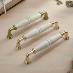 Op de afbeelding: Drie kastgrepen met witte marmerlook-middens en goudkleurige metalen hardware. De grepen hebben een gebogen ontwerp en zijn in een rij gerangschikt. De hardware heeft een klassieke, elegante stijl, geschikt voor keuken- of badkamerkasten.