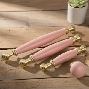 Op de afbeelding: Een set roze kastgrepen en een bijpassende knop met goudkleurige hardware. De grepen zijn in drie verschillende maten en de knop is rond. De hardware heeft een warme goudkleur. De items zijn gerangschikt op een houten oppervlak.