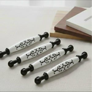 May include: Four white and black cabinet pulls with a decorative scroll design. The pulls have a ceramic center and black metal ends. The pulls are arranged diagonally on a light-colored surface.