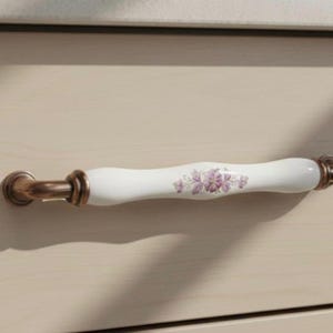 Könnte beinhalten: Ein weißer Keramik-Schrankgriff mit einem lila Blumenmuster. Die Enden sind aus bronzefarbenem Metall. Der Griff ist auf einer hellen Holzoberfläche montiert. Diese dekorative Hardware verleiht Möbeln einen Hauch von Eleganz.