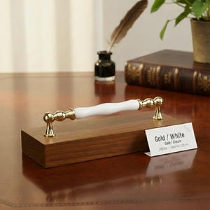 Op de afbeelding: Een goud-witte kastgreep met een wit keramisch midden en gouden accenten. De greep is gemonteerd op een houten blok. Het label luidt "Gold / White" en vermeldt afmetingen van 192 mm en 160 mm.