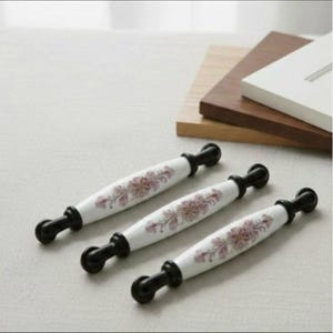 Könnte beinhalten: Drei weiße Keramik-Schrankgriffe mit einem floralen Design in Rosa und Lila. Die Griffe haben schwarze Metallenden. Die Griffe sind diagonal auf einer hellen Oberfläche angeordnet.