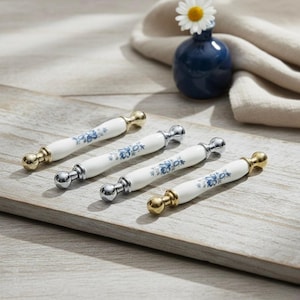 Könnte beinhalten: Vier Schrankgriffe mit weißen Keramikgriffen, blauen Blumenmustern und goldenen oder silbernen Enden. Sie sind auf einem Holzbrett angeordnet. Eine kleine blaue Vase mit einer Gänseblümchen steht im Hintergrund.