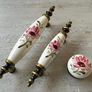 May include: A set of three decorative cabinet hardware pieces. Two white ceramic handles with pink floral designs and bronze-coloured hardware. A matching round knob with a similar floral pattern is also included.