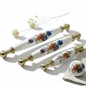 Op de afbeelding: Een set van vier witte keramische kastgrepen en een knop met een bloemenontwerp in oranje, paars en blauw. De grepen hebben goudkleurige metalen uiteinden. De knop is rond en ligt op een witte doek.